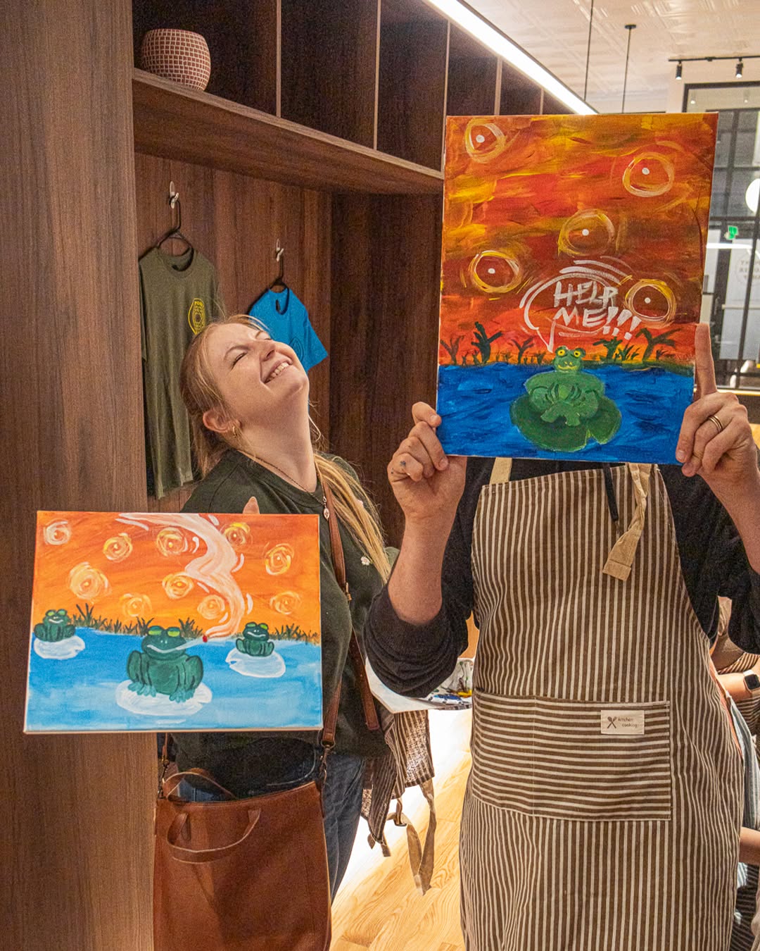 Two guests holding up their finished frog paintings at the Puff N Paint night